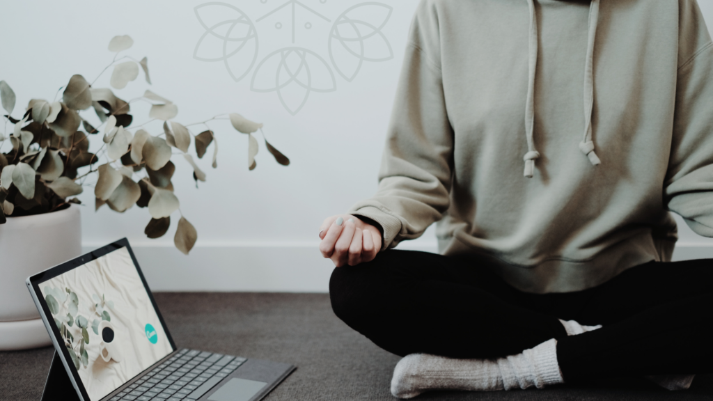 woman sitting in meditation with a soft hoodie and leggings. She has a laptop open suggesting she's listening to a guided meditation