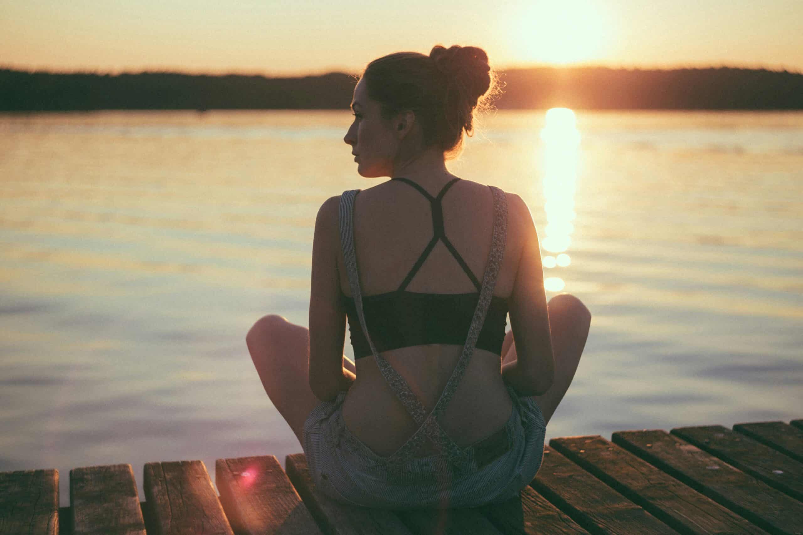 Woman meditating by the water at sunset for stress relief and cortisol reduction.