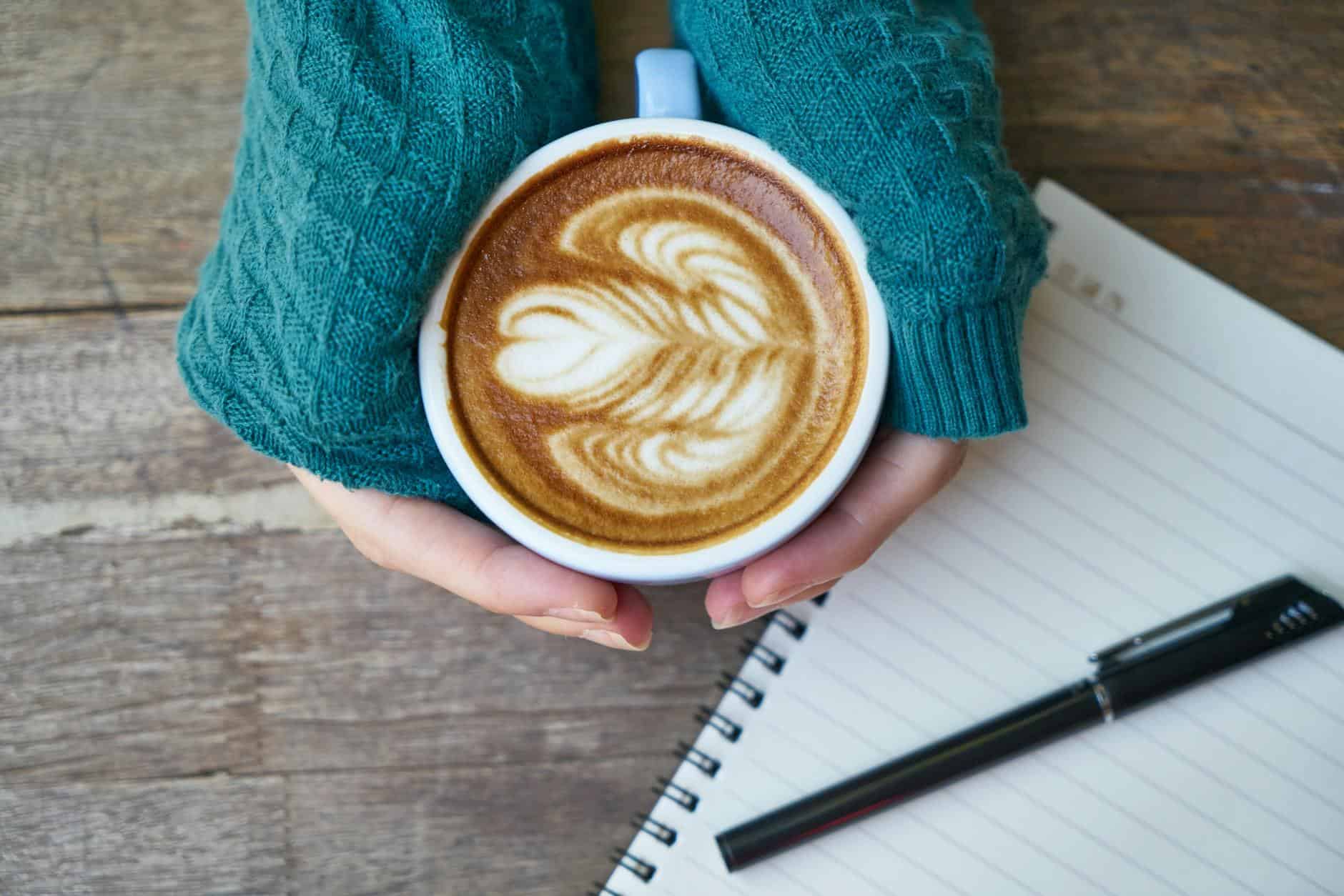 person holding cup of coffee latte near notebook and pen