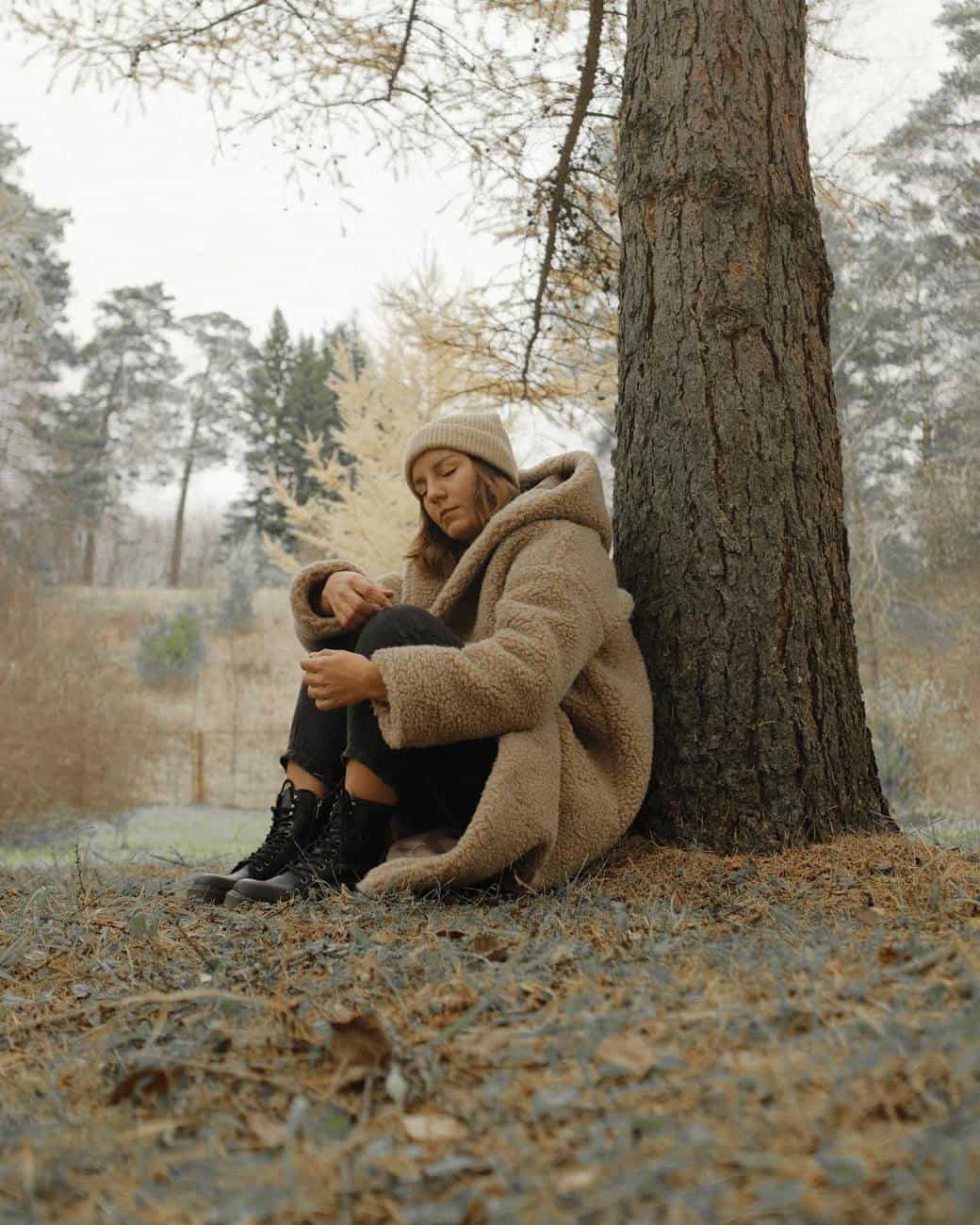 woman in beige wool coat with knit hat sitting under a tree
