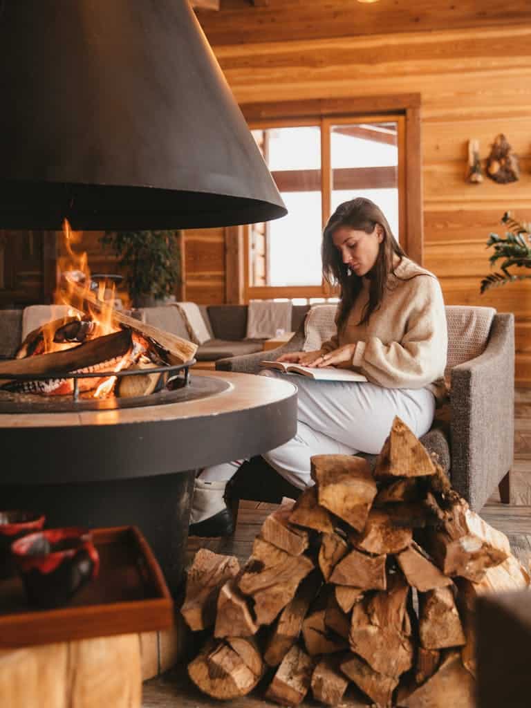 Woman reading by a warm fire with stacked logs, embracing winter indoors.