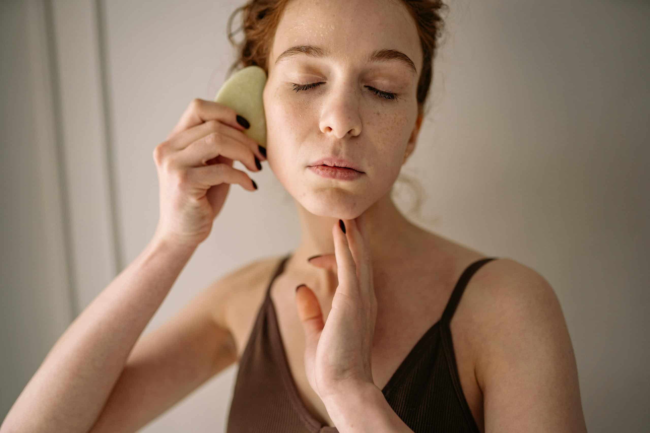 Young woman using jade stone for facial gua sha massage, focusing on self-care and skincare routine.