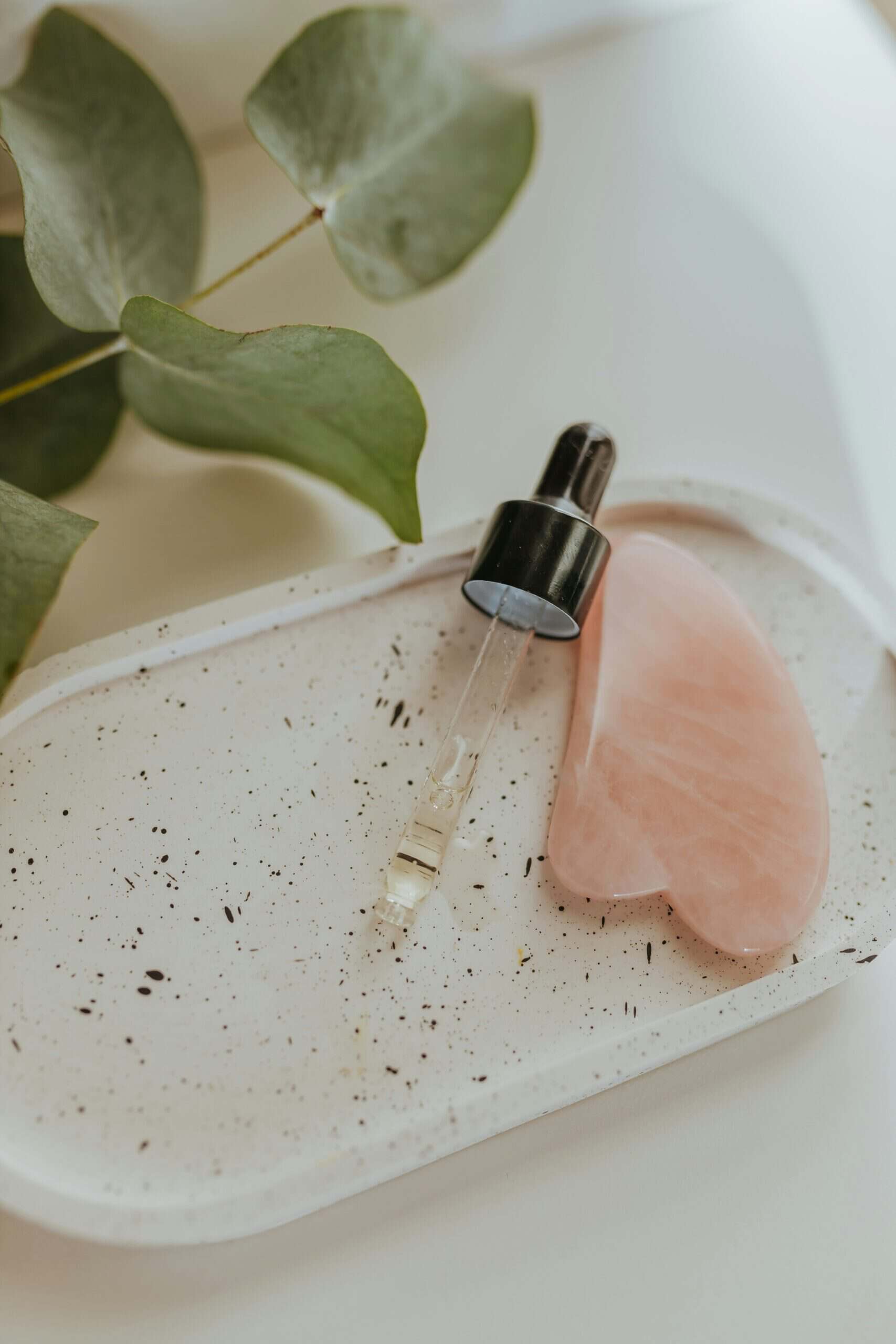 Gua Sha tool and serum dropper on a tray with eucalyptus leaves.